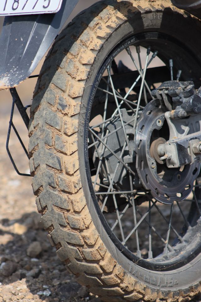 A muddy 50/50 dual-sport tyre on gravel, the kind of tread that covers most seasons reasonably well